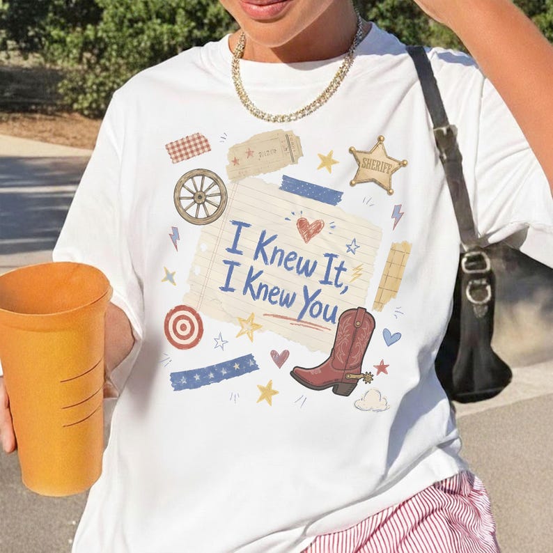 May include: A white t-shirt featuring a graphic print of a lined notebook paper with the text 'I Knew It, I Knew You' in blue script. The design includes a red cowboy boot, a sheriff star, a wagon wheel, and various small stars and hearts.