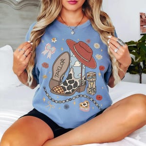 May include: A light blue crewneck sweatshirt featuring a graphic print of a cowboy boot with a cow print pattern, a red hat, alphabet blocks, and the text "TAYLOR" and "I KNEW IT I KNEW YOU" with various small floral and star accents.