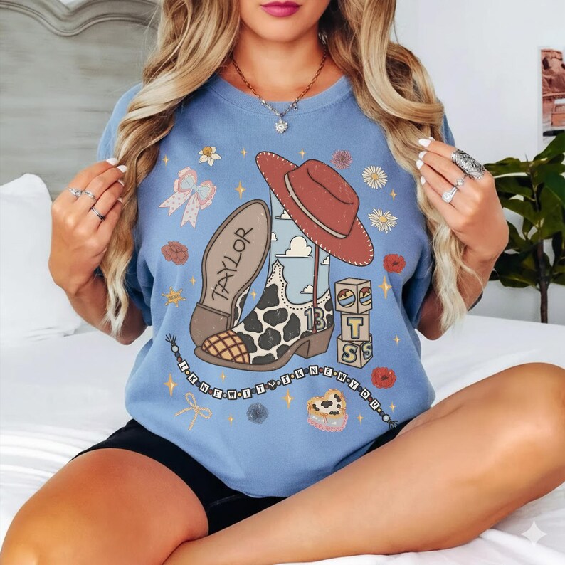 May include: A light blue crewneck sweatshirt featuring a graphic print of a cowboy boot with a cow print pattern, a red hat, alphabet blocks, and the text "TAYLOR" and "I KNEW IT I KNEW YOU" with various small floral and star accents.