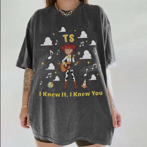 May include: A charcoal gray short-sleeve t-shirt featuring a graphic of a cowgirl playing a guitar. The design includes the text TS and I Knew It, I Knew You, surrounded by white clouds, musical notes, yellow stars, a ball, and a traffic cone.