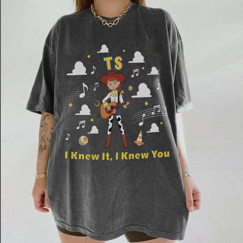 May include: A charcoal gray short-sleeve t-shirt featuring a graphic of a cowgirl playing a guitar. The design includes the text TS and I Knew It, I Knew You, surrounded by white clouds, musical notes, yellow stars, a ball, and a traffic cone.