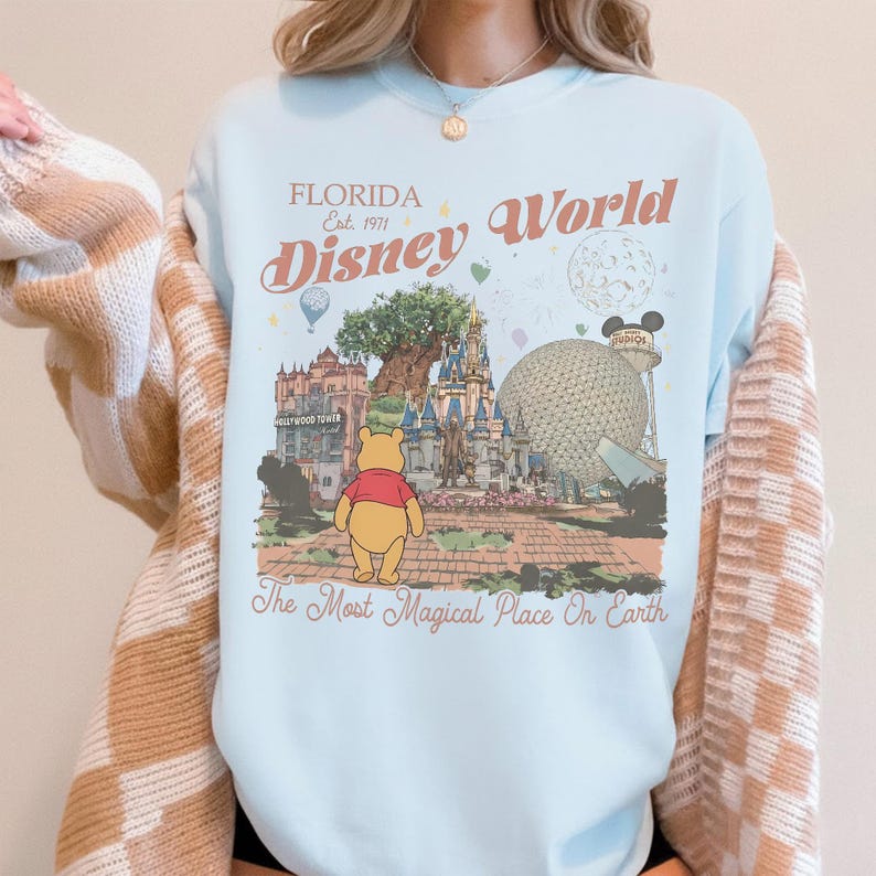Puede incluir: Sudadera azul claro con un dise&ntilde;o de Disney World en Florida. El dise&ntilde;o incluye ilustraciones del castillo, Epcot y Winnie the Pooh. El texto dice "Florida Est. 1971" y "Disney World" con la frase "The Most Magical Place On Earth".