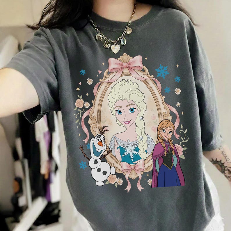 Pu&ograve; includere: Una maglietta grigio antracite con un'illustrazione incorniciata di Elsa e Anna del film Frozen, con Olaf il pupazzo di neve. L'illustrazione include un fiocco rosa, fiocchi di neve e dettagli floreali. La maglietta ha maniche corte.