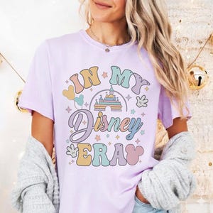 Op de afbeelding: Een lichtpaars t-shirt met de tekst "IN MY Disney ERA" in een retro lettertype. Het ontwerp bevat een kasteel, Mickey Mouse-handen en sterren in pastelkleuren. Het shirt heeft korte mouwen.