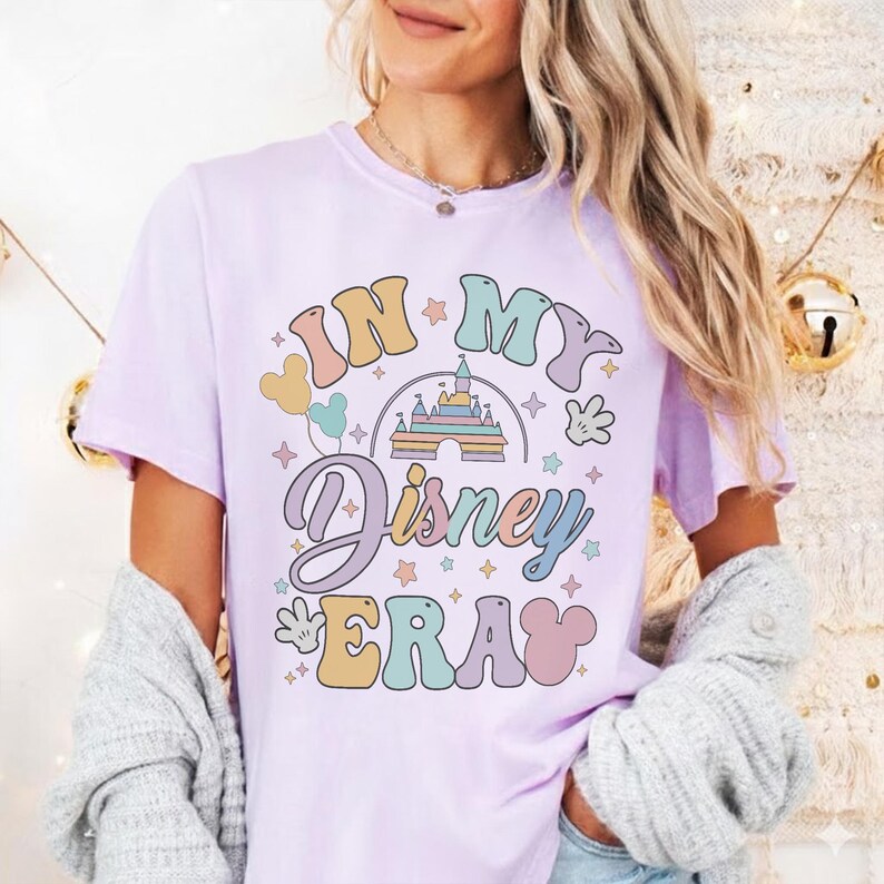 Op de afbeelding: Een lichtpaars t-shirt met de tekst "IN MY Disney ERA" in een retro lettertype. Het ontwerp bevat een kasteel, Mickey Mouse-handen en sterren in pastelkleuren. Het shirt heeft korte mouwen.