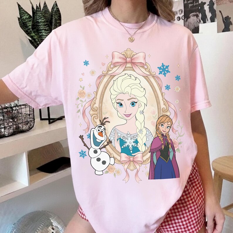 Pu&ograve; includere: T-shirt rosa chiaro con un'illustrazione a cartoni animati di Elsa, Anna e Olaf di Frozen. Il design include un ritratto di Elsa con cornice dorata, dettagli floreali e fiocchi di neve. La t-shirt &egrave; girocollo.