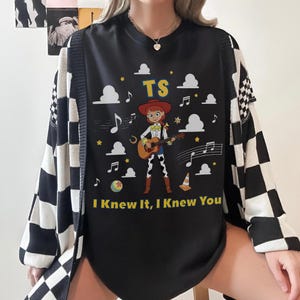 May include: A black t-shirt featuring a graphic of a cartoon cowgirl playing an acoustic guitar. The design includes white clouds, musical notes, a yellow ball, a traffic cone, and the text TS and I Knew It, I Knew You in yellow lettering.