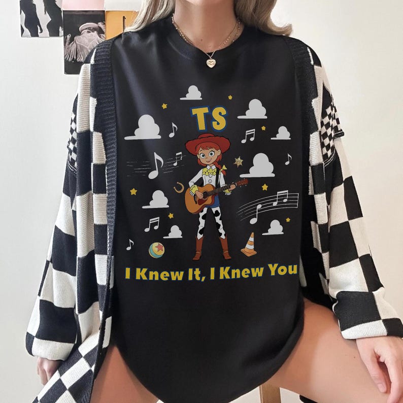 May include: A black t-shirt featuring a graphic of a cartoon cowgirl playing an acoustic guitar. The design includes white clouds, musical notes, a yellow ball, a traffic cone, and the text TS and I Knew It, I Knew You in yellow lettering.