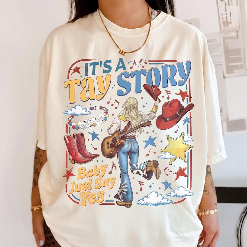 May include: An off-white t-shirt featuring a graphic print with the text IT'S A TAY STORY and Baby Just Say Yes. The design includes a person playing a guitar, red cowboy boots, a red hat, a beaded bracelet, stars, clouds, and musical notes.
