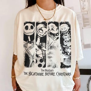 May include: A cream-colored short-sleeve t-shirt featuring a black graphic print of characters from Tim Burton's The Nightmare Before Christmas. The design includes four vertical panels displaying Jack Skellington, Sally, Oogie Boogie, and Lock, Shock, and Barrel.