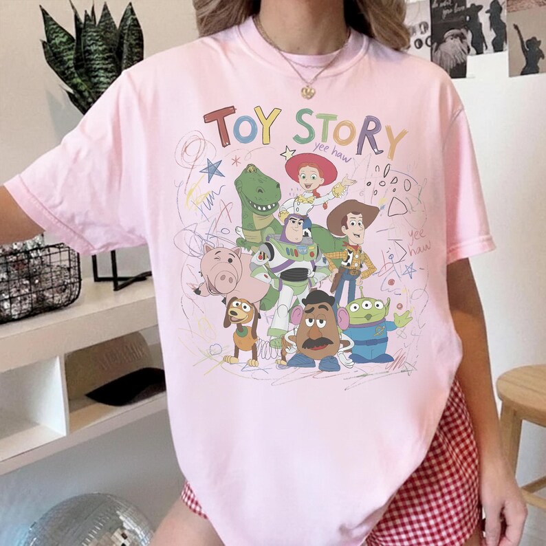 May include: A light pink t-shirt featuring the words "TOY STORY" in colorful letters above a graphic of characters from the animated film. The design includes Woody, Buzz Lightyear, and other characters. The shirt is a crew neck.