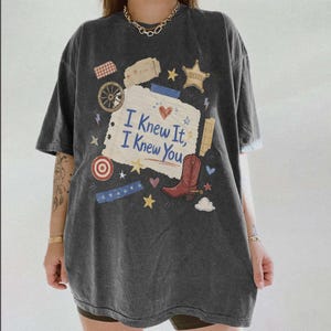 May include: A charcoal gray short-sleeve t-shirt featuring a central graphic of a torn piece of paper with the text "I Knew It, I Knew You" in blue. The design includes a red cowboy boot, a sheriff star, a wagon wheel, and various small stars.