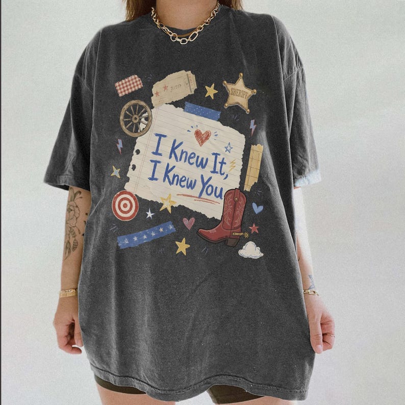 May include: A charcoal gray short-sleeve t-shirt featuring a central graphic of a torn piece of paper with the text "I Knew It, I Knew You" in blue. The design includes a red cowboy boot, a sheriff star, a wagon wheel, and various small stars.