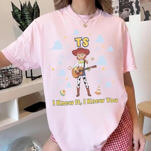 May include: A light pink t-shirt featuring a graphic of a cartoon cowgirl playing an acoustic guitar. The design includes the text TS above the character and I Knew It, I Knew You below, surrounded by small blue clouds, stars, and musical notes.