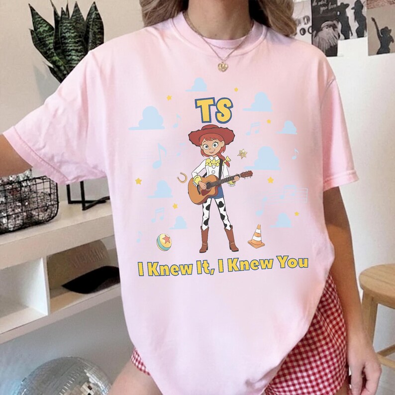 May include: A light pink t-shirt featuring a graphic of a cartoon cowgirl playing an acoustic guitar. The design includes the text TS above the character and I Knew It, I Knew You below, surrounded by small blue clouds, stars, and musical notes.