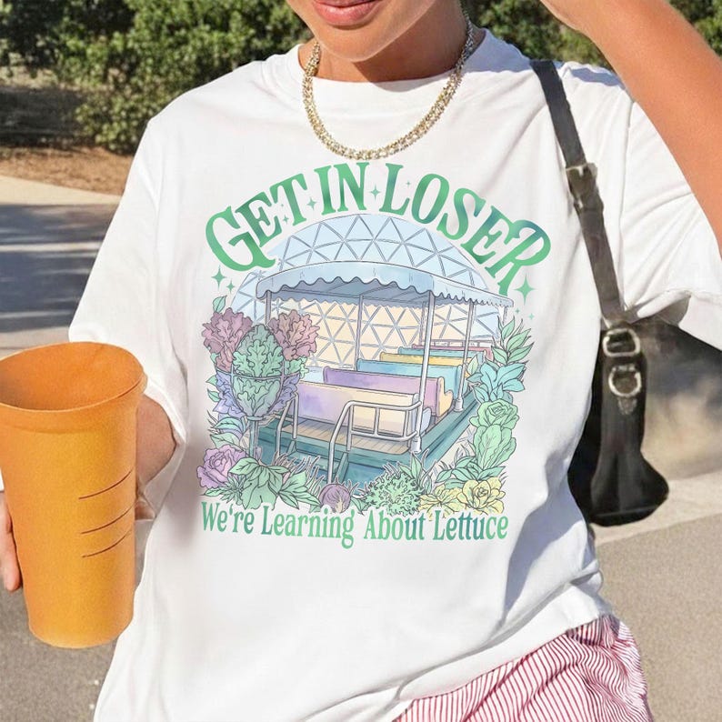 Puede incluir: Camiseta blanca con un gr&aacute;fico colorido que presenta una atracci&oacute;n de parque de atracciones caprichosa y el texto "GET IN LOSER" encima de "We're Learning About Lettuce". El dise&ntilde;o incluye colores pastel y elementos florales.
