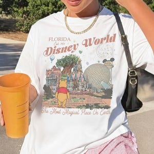 Puede incluir: Camiseta blanca con un gr&aacute;fico colorido de Disney World en Florida. El dise&ntilde;o incluye el texto "Disney World" y "The Most Magical Place On Earth", junto con im&aacute;genes de lugares emblem&aacute;ticos del parque y Winnie the Pooh.