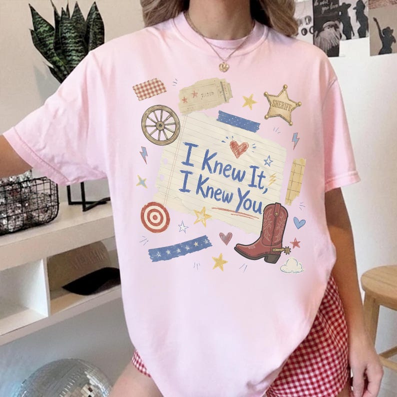 May include: A light pink short-sleeve t-shirt featuring a central graphic on a lined paper background with the text I Knew It, I Knew You. The design includes a brown cowboy boot, a sheriff star, a wagon wheel, and various small colorful icons.