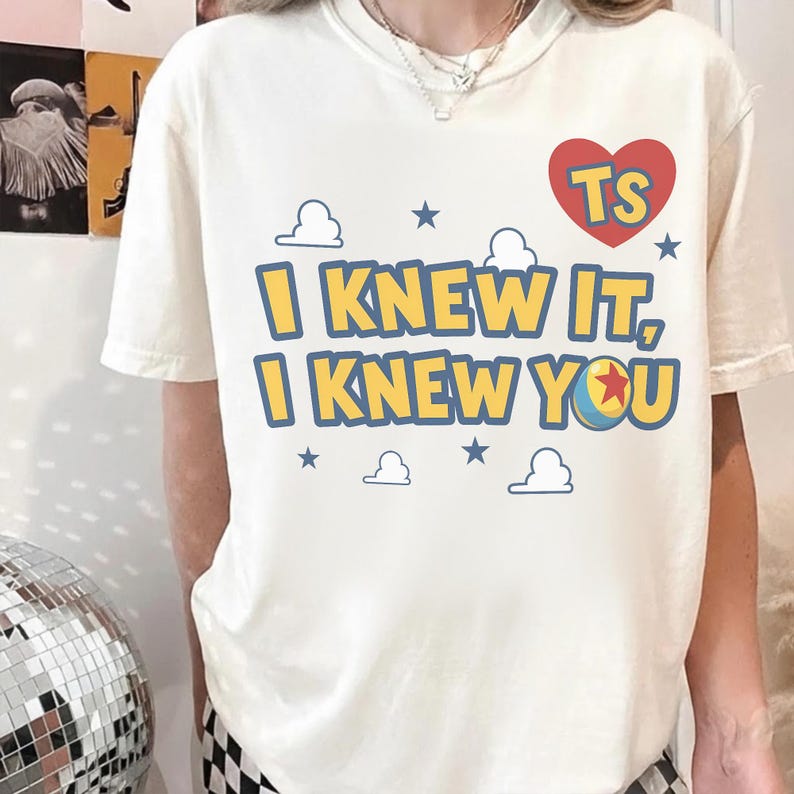 May include: A cream-colored short-sleeve t-shirt featuring a graphic print with the text "I KNEW IT, I KNEW YOU" in yellow letters with blue outlines. The design includes a red heart with "TS" inside, blue stars, and white cloud outlines.