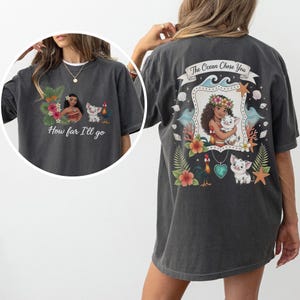 May include: Dark grey t-shirt with a colourful graphic of a woman, a pig, and a rooster. The front of the shirt has the text "How far I'll go". The back of the shirt has the text "The Ocean Chose You".