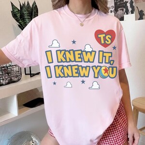 May include: A light pink short-sleeve t-shirt featuring a graphic print with the text "I KNEW IT, I KNEW YOU" in yellow letters with blue outlines. The design includes a red heart with "TS" inside, small blue stars, white clouds, and a ball.