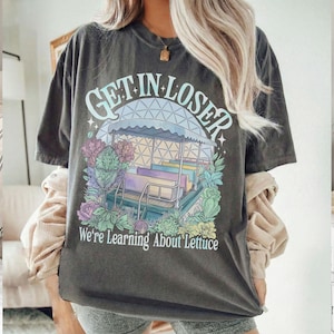 Puede incluir: Camiseta gris oscuro con el texto "GET IN LOSER" sobre un gr&aacute;fico colorido de un paseo y el texto "We're Learning About Lettuce" debajo. La camiseta tiene un aspecto vintage y desgastado.