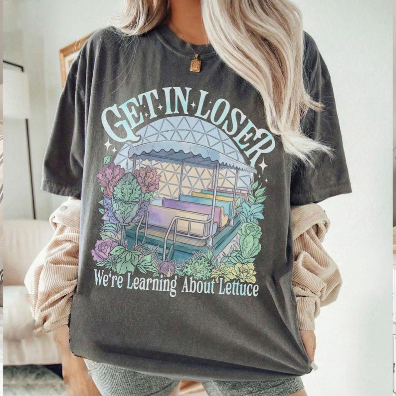 Puede incluir: Camiseta gris oscuro con el texto "GET IN LOSER" sobre un gr&aacute;fico colorido de un paseo y el texto "We're Learning About Lettuce" debajo. La camiseta tiene un aspecto vintage y desgastado.