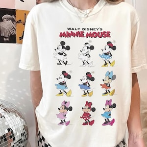K&ouml;nnte beinhalten: Cremefarbenes T-Shirt mit dem Text "WALT DISNEY'S MINNIE MOUSE" in Rot und Schwarz. Das Shirt zeigt neun verschiedene Minnie Mouse-Illustrationen, jede in einem einzigartigen Outfit, mit verschiedenen farbigen R&ouml;cken und Schleifen.