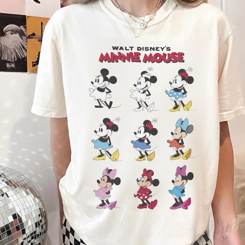 K&ouml;nnte beinhalten: Cremefarbenes T-Shirt mit dem Text "WALT DISNEY'S MINNIE MOUSE" in Rot und Schwarz. Das Shirt zeigt neun verschiedene Minnie Mouse-Illustrationen, jede in einem einzigartigen Outfit, mit verschiedenen farbigen R&ouml;cken und Schleifen.