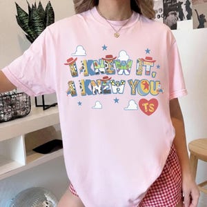 May include: A light pink short-sleeve t-shirt featuring a graphic print with the text I KNEW IT, I KNEW YOU and TS inside a red heart. The letters are decorated with patterns inspired by toy characters, including cowboy hats, clouds, and stars.