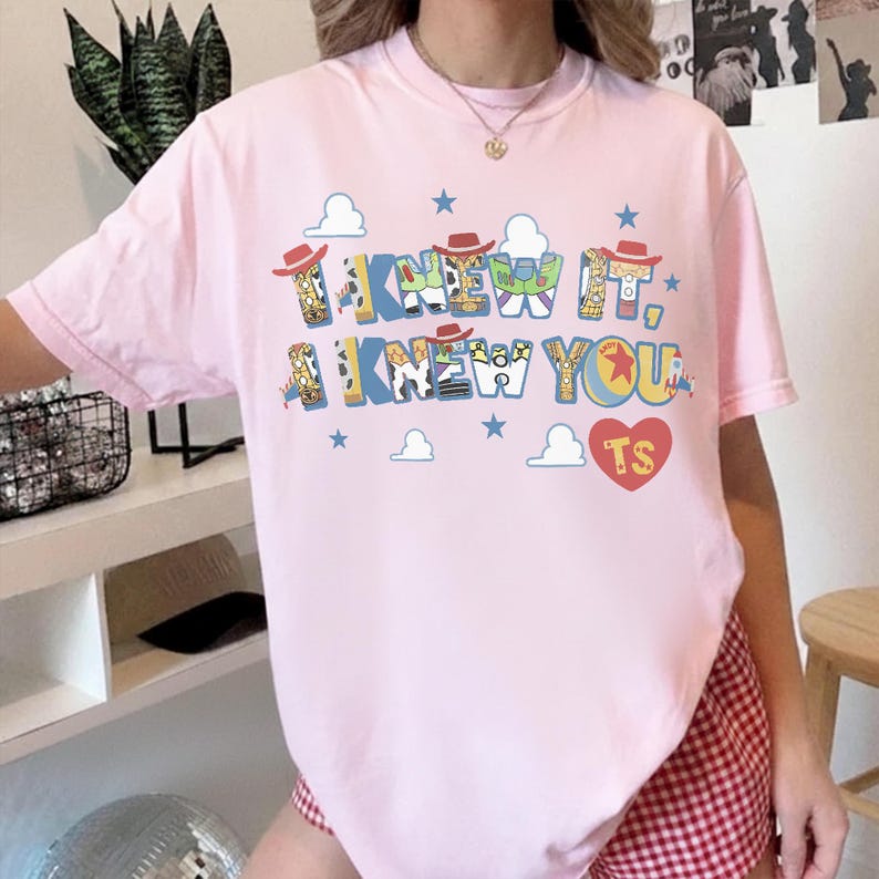 May include: A light pink short-sleeve t-shirt featuring a graphic print with the text I KNEW IT, I KNEW YOU and TS inside a red heart. The letters are decorated with patterns inspired by toy characters, including cowboy hats, clouds, and stars.