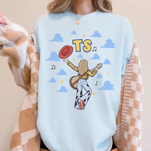 May include: A light blue sweatshirt featuring a graphic illustration of a person with long hair wearing a yellow plaid shirt and cow-print bell-bottom pants. The person holds a guitar and a red hat, with the text TS and blue clouds in the background.