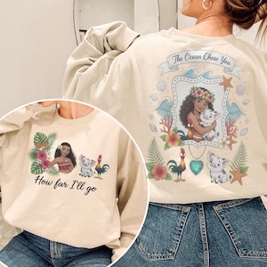 May include: Cream-coloured sweatshirt with a vibrant graphic of a woman with a flower crown, holding a pig, and the text "The Ocean Chose You". The front has the text "How far I'll go" and a matching graphic.