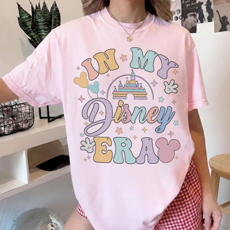 Op de afbeelding: Een lichtroze t-shirt met de tekst "IN MY Disney ERA" in pastelkleuren. Het ontwerp bevat een kasteel, Mickey Mouse-handschoenen en sterren. Het is een casual kledingstuk.