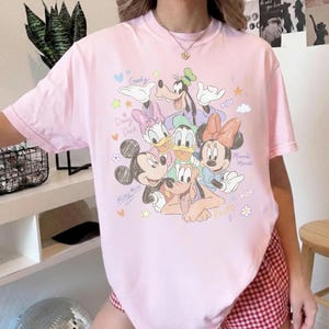 May include: Pink t-shirt featuring a colorful graphic of Disney characters including Mickey Mouse, Minnie Mouse, Goofy, Donald Duck, Daisy Duck, and Pluto. The shirt has the characters' names and other decorative elements.