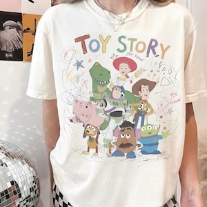 May include: Cream-colored t-shirt featuring a colorful graphic of characters from the animated film "Toy Story." The design includes the words "Toy Story" in a playful font, along with drawings and the phrase "yee haw."