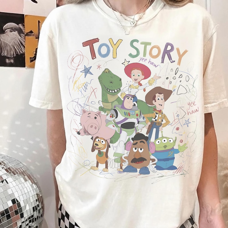 May include: Cream-colored t-shirt featuring a colorful graphic of characters from the animated film "Toy Story." The design includes the words "Toy Story" in a playful font, along with drawings and the phrase "yee haw."