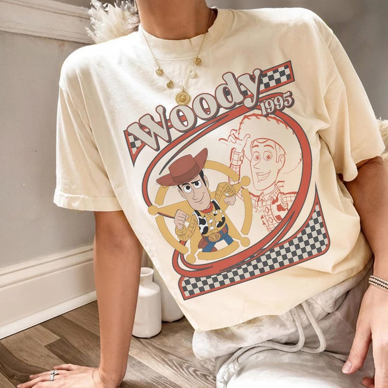 May include: Cream-colored t-shirt featuring a graphic of Woody from Toy Story. The design includes Woody's image, the word "Woody" in a retro font, and the year "1995". The shirt has short sleeves and a relaxed fit.