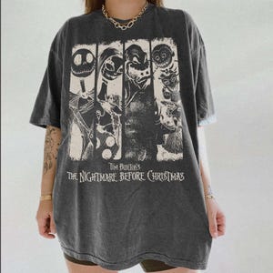 May include: A charcoal gray short-sleeve t-shirt featuring a graphic print of characters from Tim Burton's The Nightmare Before Christmas. The design includes four vertical panels with illustrations of Jack Skellington, Sally, and other figures.
