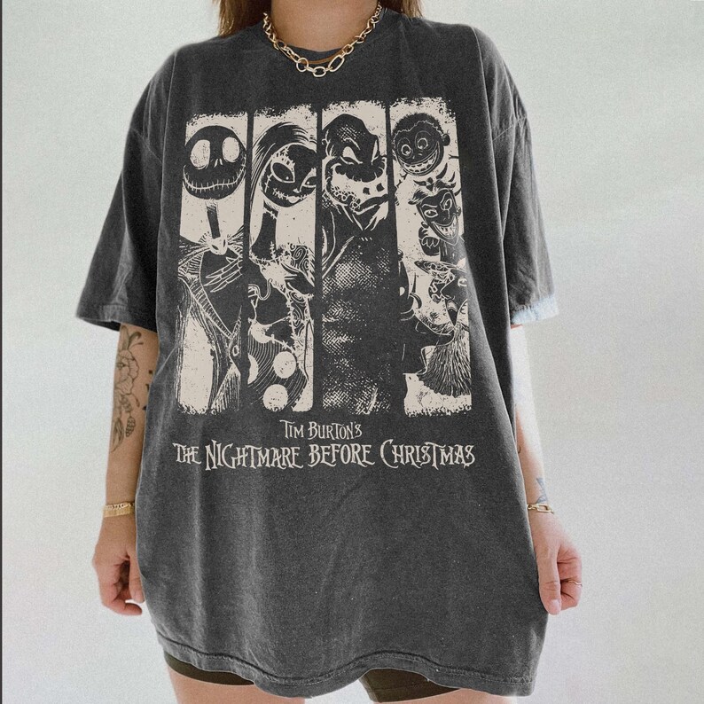 May include: A charcoal gray short-sleeve t-shirt featuring a graphic print of characters from Tim Burton's The Nightmare Before Christmas. The design includes four vertical panels with illustrations of Jack Skellington, Sally, and other figures.