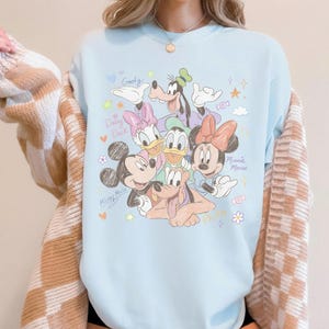 May include: Light blue crew neck sweatshirt featuring a colorful graphic of Disney characters Mickey Mouse, Minnie Mouse, Donald Duck, Daisy Duck, Goofy, and Pluto. The names of the characters are written in a playful font. The sweatshirt has a relaxed fit.
