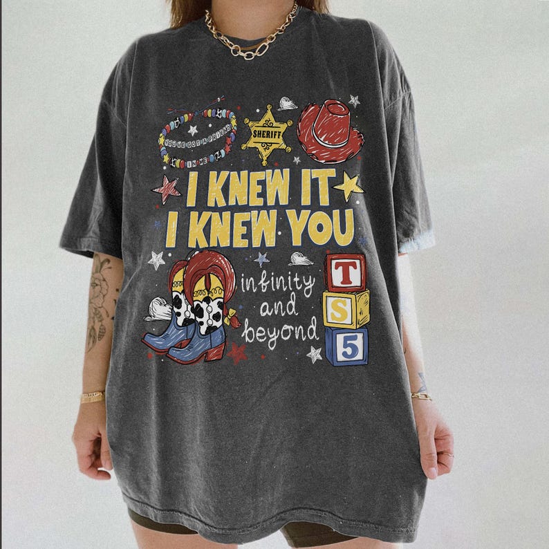 May include: A charcoal gray short-sleeve t-shirt featuring a graphic print with the text I KNEW IT I KNEW YOU infinity and beyond. The design includes a sheriff star, a red cowboy hat, cow-print boots, and three stacked blocks labeled T S 5.