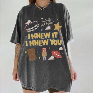 May include: A charcoal grey short-sleeve t-shirt featuring a graphic print with the text "I KNEW IT I KNEW YOU", "infinity and beyond", and "YOU'VE GOT A FRIEND IN ME". The design includes a sheriff badge, cowboy boot, hat, and blocks.