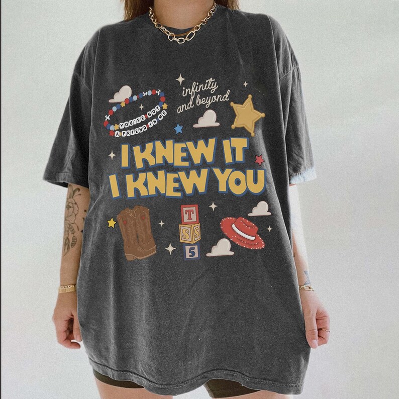 May include: A charcoal grey short-sleeve t-shirt featuring a graphic print with the text "I KNEW IT I KNEW YOU", "infinity and beyond", and "YOU'VE GOT A FRIEND IN ME". The design includes a sheriff badge, cowboy boot, hat, and blocks.