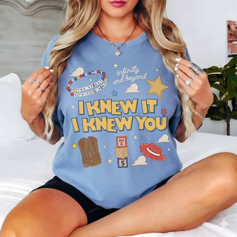 May include: A light blue crewneck t-shirt featuring a graphic print with the text "I KNEW IT I KNEW YOU", "infinity and beyond", and "YOU'VE GOT A FRIEND IN ME". The design includes illustrations of cowboy boots, a red hat, and alphabet blocks.