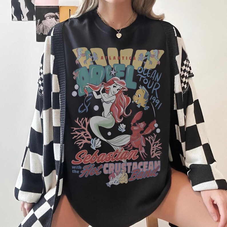 May include: Black graphic tee featuring Ariel from The Little Mermaid, with the text "Atlantica Hotel Ocean Tour 1991". The tee is paired with a black and white checkered cardigan.