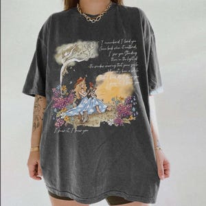 May include: A charcoal gray oversized t-shirt featuring a graphic print of a person in a blue dress and cowboy boots sitting in a field of flowers. The design includes handwritten-style text that reads, I remembered I loved you, and more.