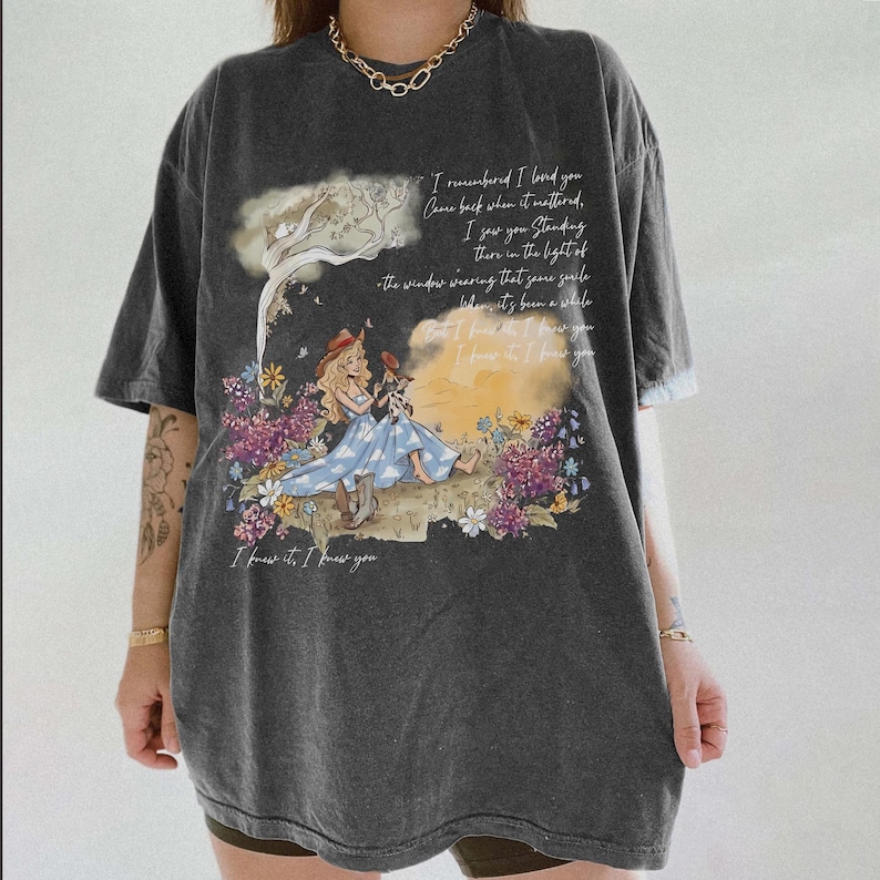 May include: A charcoal gray oversized t-shirt featuring a graphic print of a person in a blue dress and cowboy boots sitting in a field of flowers. The design includes handwritten-style text that reads, I remembered I loved you, and more.