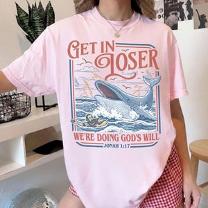 K&ouml;nnte beinhalten: Hellrosa T-Shirt mit einem Grafikdesign, das einen Wal, Wellen und eine Person im Wasser zeigt. Der Text auf dem Shirt lautet "Get in Loser" &uuml;ber dem Wal und "We're Doing God's Will" darunter, mit "Jonah 1:17" unten.