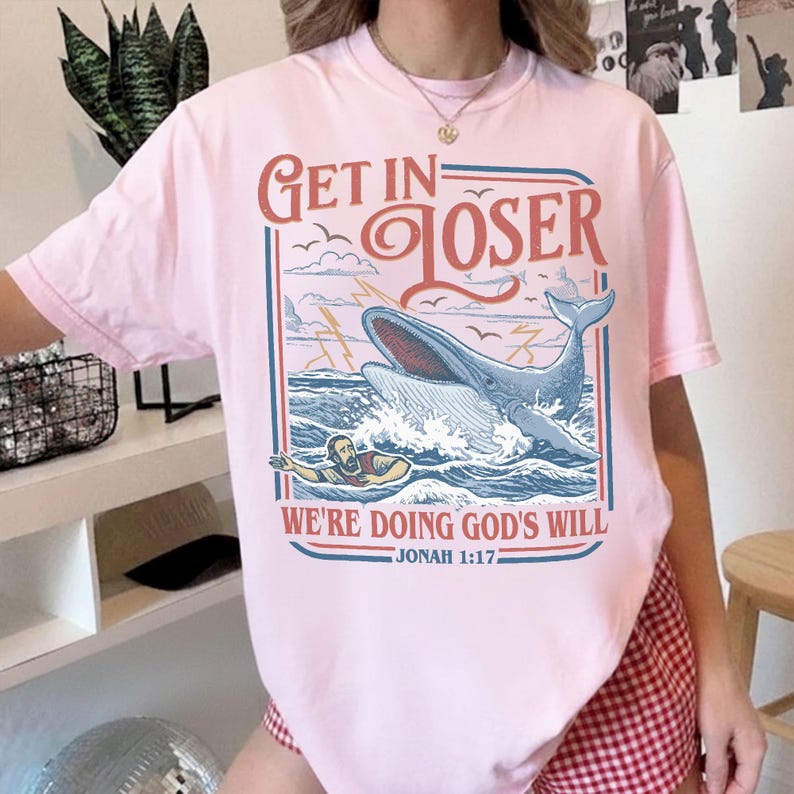 K&ouml;nnte beinhalten: Hellrosa T-Shirt mit einem Grafikdesign, das einen Wal, Wellen und eine Person im Wasser zeigt. Der Text auf dem Shirt lautet "Get in Loser" &uuml;ber dem Wal und "We're Doing God's Will" darunter, mit "Jonah 1:17" unten.